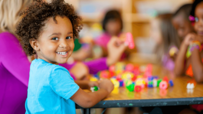 A little girl looking back at the camera smiling while playing with blocks on a table with a group.